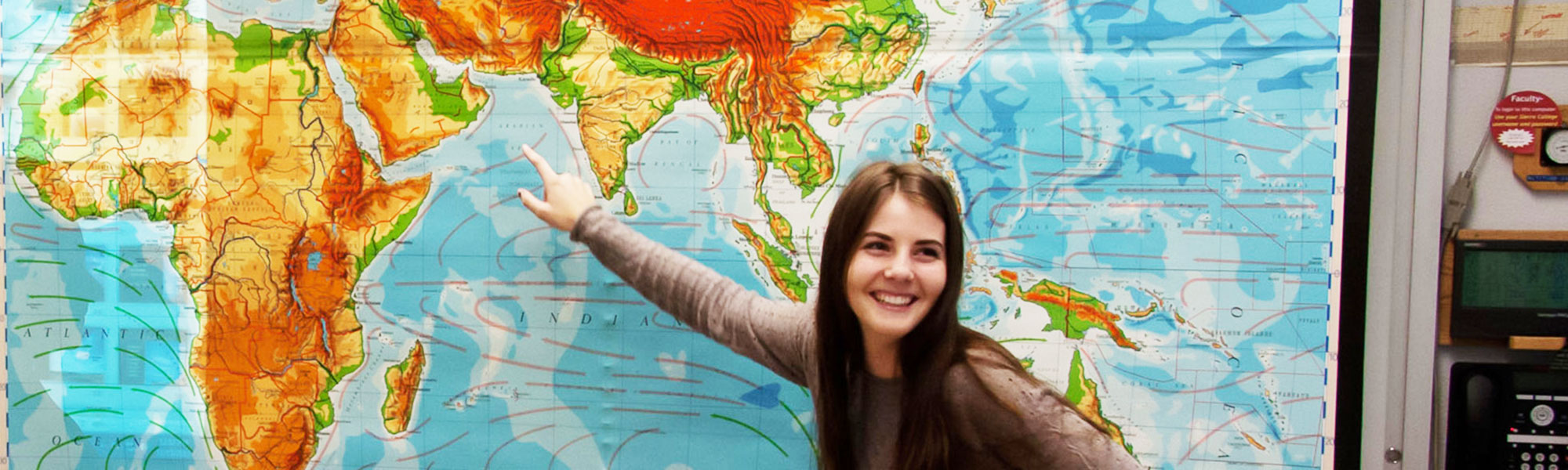 Female student pointing at world map in front of classroom