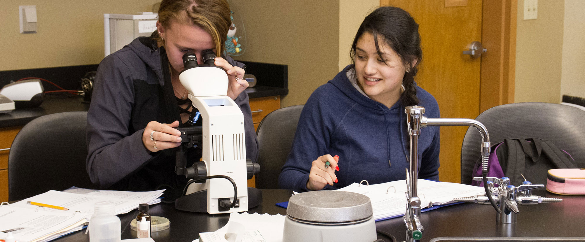 Female biology student uses microscope as her partner writes down data.